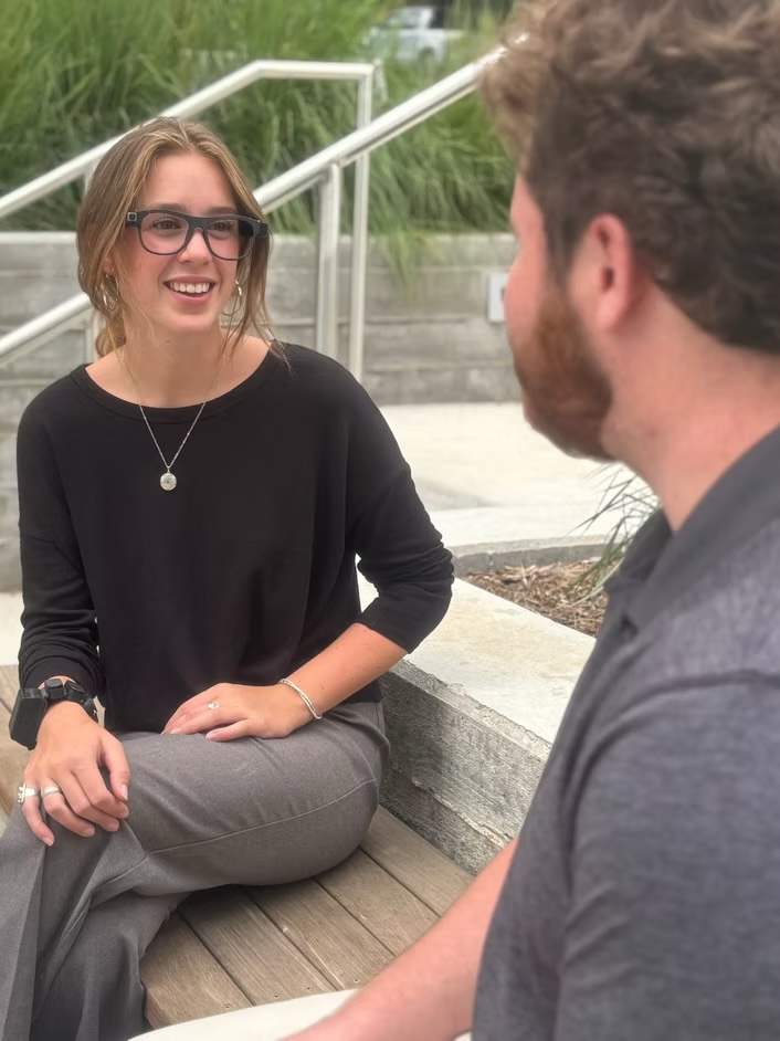 Two people seated, facing each other in conversation, with one person wearing the AlEye wristband and glasses, smiling at the other.